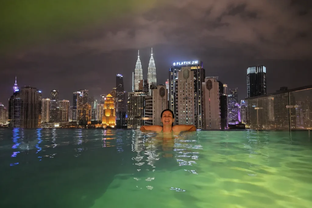 Swimmer in a rooftop infinity pool overlooking the illuminated Petronas Towers skyline at night, Kuala Lumpur