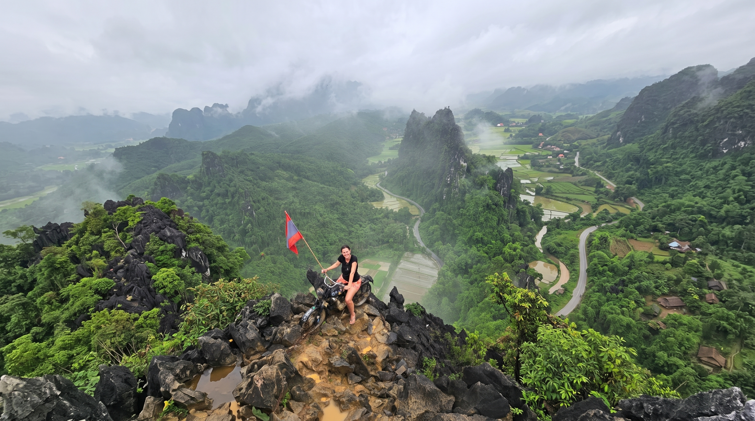 Vang Vieng travel guide: traveler holding a flag on a motorbike atop a rocky summit overlooking the karst valley and rice paddies