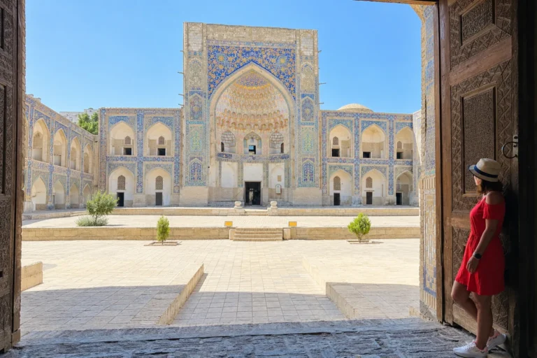 Bukhara travel guide: traveler in a red dress and sun hat looking at a tiled madrasah courtyard